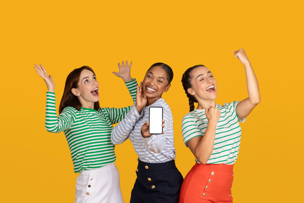 Emotional young ladies girlfriends showing smartphone with white blank screen mockup copy space gesturing and exclaiming, celebrating beginning of season sale, yellow background - Фото, зображення