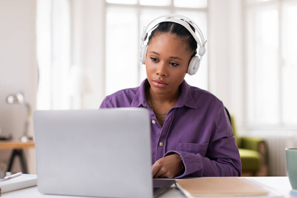 Mujer joven enfocada que usa auriculares mientras trabaja en su computadora portátil, vestida con camisa púrpura, profundamente concentrada en su tarea de aprendizaje electrónico en casa - Foto, Imagen