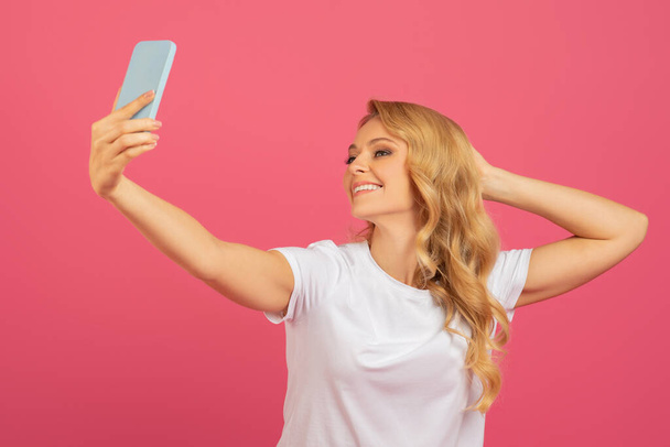 Young attractive blonde taking a selfie with her smartphone, posing for photo in casual white t-shirt, standing on pink studio backdrop. Concept of social media and online presence - Photo, Image