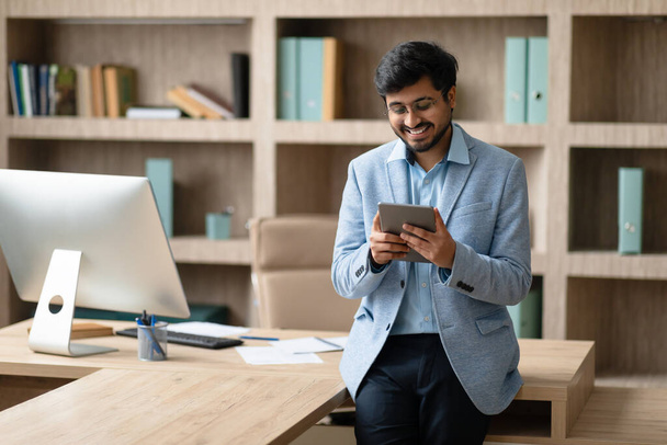 Веселый индийский профессиональный бизнесмен веб-серфинг на цифровом планшете во время работы в современной офисной среде, просматривая веб на современном рабочем месте. Интернет и технологии для бизнеса - Фото, изображение
