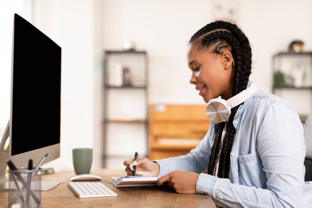 Engagierte schwarze Studentin mit Kopfhörern schreibt aufmerksam Notizen von ihrem Computer, die auf die aktive Teilnahme an einem Online-Kurs hinweisen - Foto, Bild