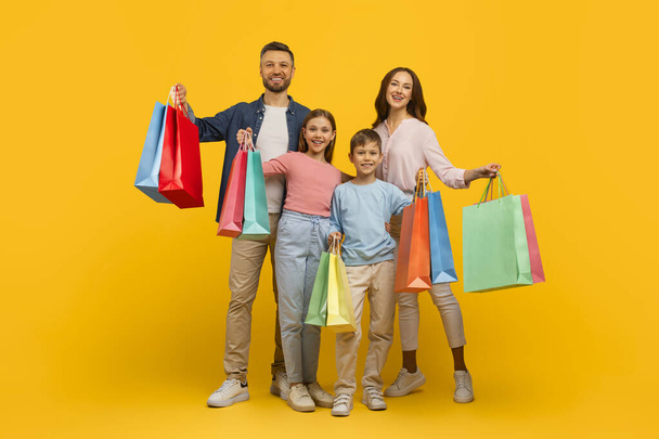 Rodzinne zakupy. Radosni młodzi rodzice i ich dzieci trzymają jasne torby na zakupy i bawią się razem stojąc na żółtym tle, radosna mama, tata i dzieci korzystające z sezonowej sprzedaży - Zdjęcie, obraz