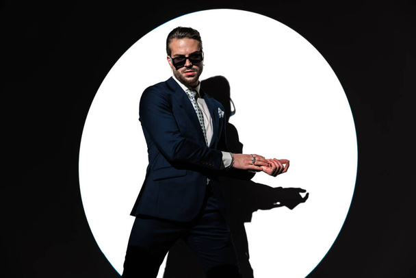 attraktiver unrasierter Mann im Anzug mit Sonnenbrille, der die Handflächen zur Seite reibt und vor Scheinwerferhintergrund posiert - Foto, Bild