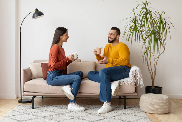 Ευτυχισμένοι νέοι σύζυγοι κουβεντιάζοντας και πίνοντας καφέ μαζί στο σπίτι, αγαπώντας Millennial ζευγάρι χαλαρώνοντας σε άνετο καναπέ στο σαλόνι, μιλώντας και χαμογελώντας, απολαμβάνοντας το χρόνο με τον άλλο - Φωτογραφία, εικόνα