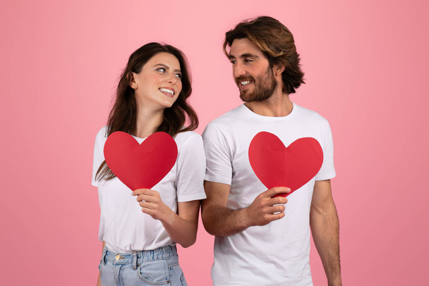 Mujer y hombre europeos sonrientes sosteniendo cada uno un corazón de papel rojo sobre sus camisetas blancas, mirándose con afecto, simbolizando el amor y el día de San Valentín sobre un fondo rosa - Foto, imagen