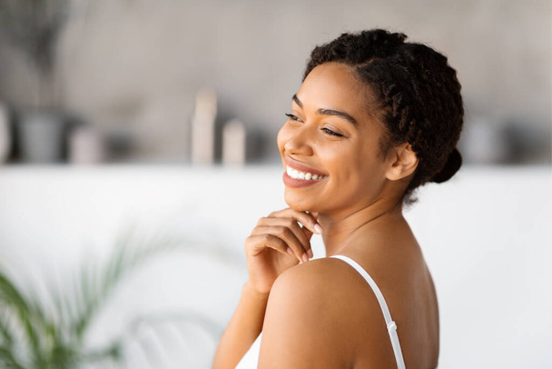 Portrait d'une jeune femme noire heureuse portant un débardeur blanc au menton souriant et touchant, charmante femme afro-américaine posant dans l'intérieur de la salle de bain, profitant de la routine beauté à la maison, espace libre - Photo, image