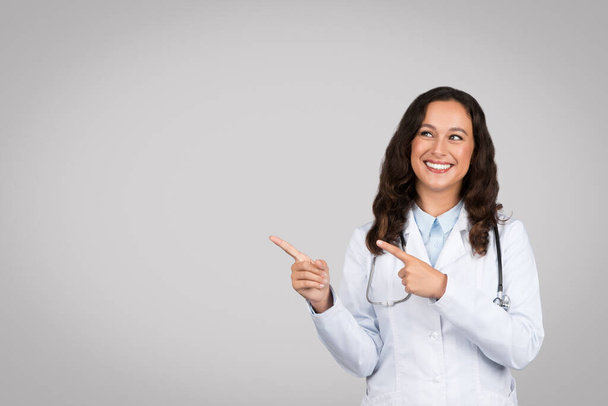 Счастливая барышня-врач в белом пальто, указывая пальцем на свободное пространство, расклеивает медицинскую рекламу и предложение на сером студийном фоне. Профессиональные консультации, здравоохранение - Фото, изображение