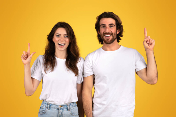 Uma mulher europeia alegre e um homem com barbas apontam para cima com sorrisos brilhantes, vestindo camisetas brancas, em pé junto contra um fundo amarelo com vibrações positivas, estúdio - Foto, Imagem