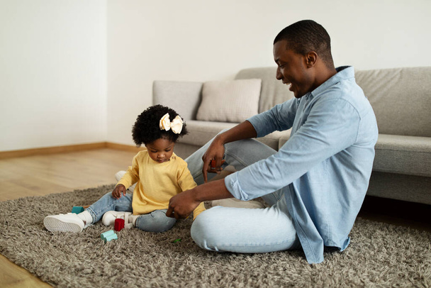 Čas na hraní. Veselý milující africký americký otec hraje barevné hračky se svou dcerou, dívka pohybující kostky, tráví volný čas spolu doma interiéru, kopírovat prostor - Fotografie, Obrázek