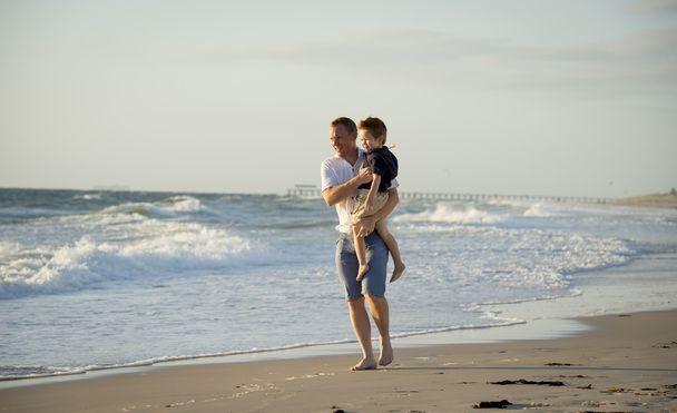 young happy father holding in his arms little son walking on beach sand in front of sea waves - Photo, Image
