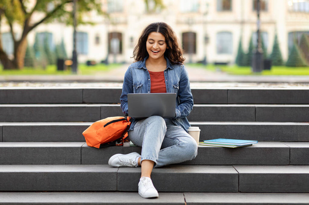 Iloinen nuori intialainen nainen opiskelija opiskelee verkossa, käyttää kannettavaa tietokonetta ulkona, istuu portailla yliopiston kampuksella, tekee kotitehtäviä, kopioi tilaa. Verkko-oppimisen käsite - Valokuva, kuva