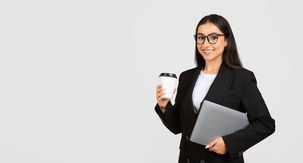 Αυτοπεποίθηση νεαρή επιχειρηματίας κρατώντας takeaway φλιτζάνι καφέ και φορητό υπολογιστή, έτοιμο για εργασία, στέκεται πάνω από γκρι φόντο, πανόραμα με χώρο αντίγραφο - Φωτογραφία, εικόνα