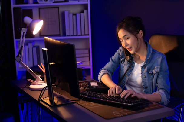 Fiatal gyönyörű ázsiai kreatív nő hívja a telefon íróasztal, hogy az ügyfél vagy munkatárs elmagyarázza alkotói projektek nagy profit, miközben keres a piaci elemzés a pc neon irodában éjszaka. Stratagem. - Fotó, kép
