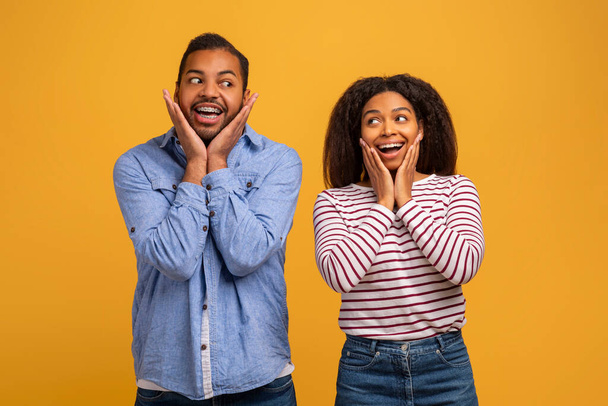 Amazed young black couple with hands on cheeks showing surprised expressions, cheerful african american man and woman standing wide-eyed and excited against yellow studio background, copy space - Photo, Image
