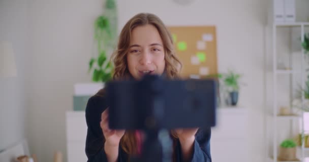Αργή κίνηση χαμογελαστή νεαρή γυναίκα δημιουργός περιεχομένου vlogging μέσω κινητού τηλεφώνου στο τρίποδο στο σπίτι - Πλάνα, βίντεο