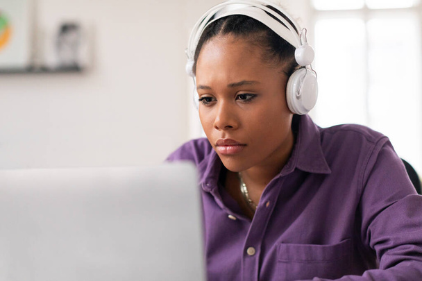 Odaklanmış siyah kız öğrenci bilgisayar ekranına odaklanıyor, beyaz kulaklık takarak derslerine dalmış, muhtemelen internetten öğreniyor. - Fotoğraf, Görsel