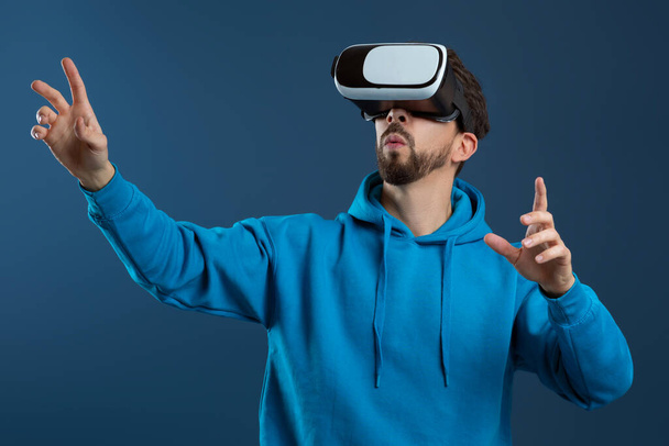 Verstrickter Mann mit Virtual-Reality-Headset und gestikulierenden Händen, verblüffter Millennial-Typ, der mit der digitalen Welt interagiert, moderne Technologien genießt, vor blauem Studiohintergrund steht - Foto, Bild