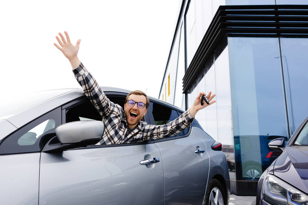 Ενθουσιασμένος νεαρός άνδρας που δείχνει ένα κλειδί αυτοκινήτου, κάθεται μέσα στο νέο του όχημα. - Φωτογραφία, εικόνα