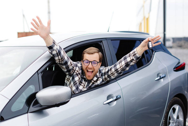 Jeune homme excité montrant une clé de voiture, assis à l'intérieur de son nouveau véhicule. - Photo, image
