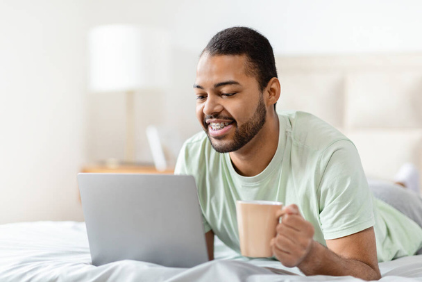 Szczęśliwy, pogodny, przystojny, afrykański mężczyzna w piżamie chłodzący się na łóżku, korzystający z laptopa, korzystający z weekendu w domu, websurfing, rozmawiający z paniami, pijący kawę, przestrzeń do kopiowania - Zdjęcie, obraz