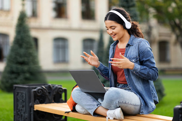 Cute young eastern woman student sitting on bench, using computer laptop and wireless headphones outdoors at university campus, have online lesson, attending webinar, copy space - Photo, Image