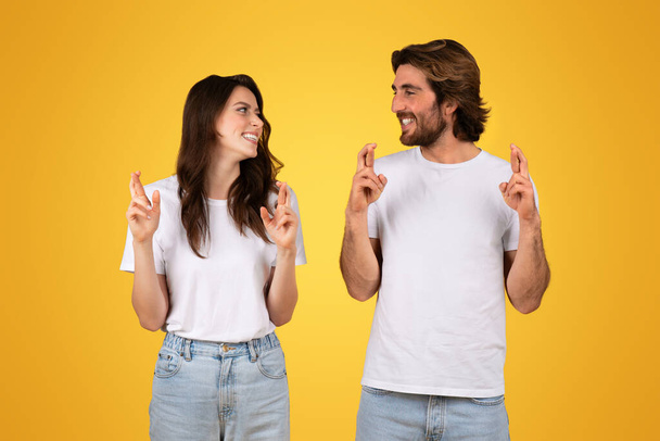 Оптимістична усміхнена кавказька молода пара в білих футболках, що перетинають пальці на удачу, дивлячись один на одного з сподіваними посмішками на жовтому тлі, студія - Фото, зображення