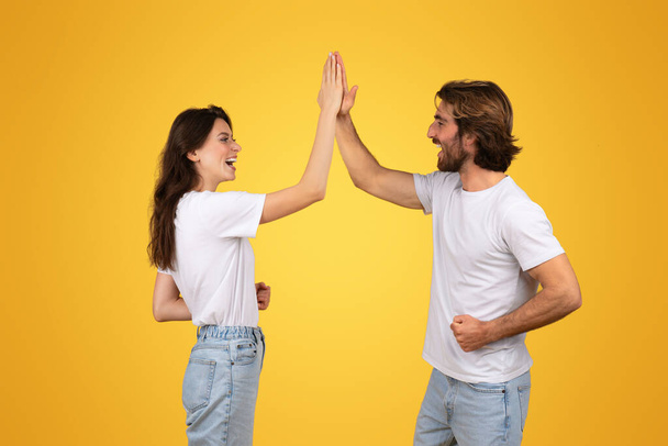 Joyful european man and woman in white shirts high-fiving, showcasing teamwork and excitement, with wide smiles and denim jeans against a monochrome yellow backdrop, studio - Photo, Image