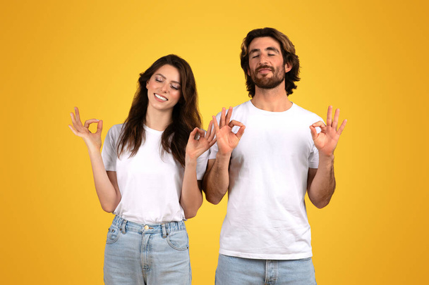 Homem e mulher europeus com olhos fechados fazendo sinais OK, sinalizando aprovação e satisfação em confortáveis camisetas brancas e jeans contra um fundo amarelo ensolarado - Foto, Imagem