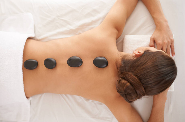 Hot stone, massage and top view of woman in salon to relax for skincare, beauty or pamper body for wellness at luxury spa. Therapy, above and person with rocks in treatment for back, spine and health. - Photo, Image