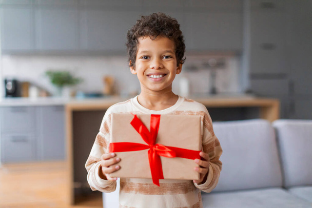 Boldog kis fekete fiú büszkén tartja csomagolt ajándék élénk piros szalaggal, aranyos tizenéves fiú mosolyog a kamera, készen áll, hogy ajándékot, áll a modern, jól megvilágított nappali belső, másolás tér - Fotó, kép