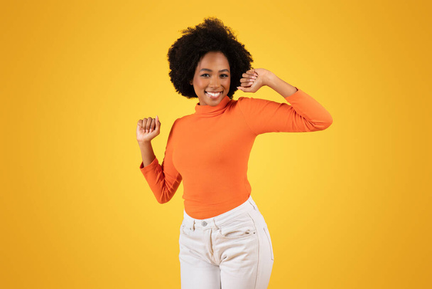 Radostná mladá Afroameričanka s hravým mrknutím oka, tančící v oranžovém roláku a bílých kalhotách na zářivém žlutém pozadí, studio. Zábava, volný čas a relaxace - Fotografie, Obrázek