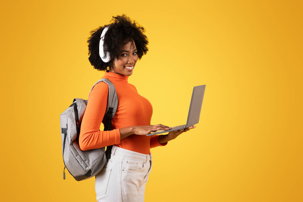 Smiling young African American woman with headphones using a laptop, wearing a casual orange turtleneck, with a gray backpack over her shoulder on a yellow background, studio - Photo, Image