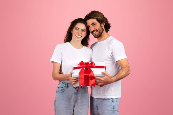 Улыбающаяся кавказская молодая пара в повседневных белых футболках, разделяющая особенный момент, с мужчиной, дарящим белый подарочный коробок с яркой красной лентой своему партнеру на розовом фоне. - Фото, изображение