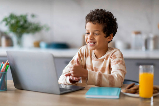 Glücklicher schwarzer Junge interagiert mit Laptop, während er am Tisch in der Küche sitzt, mit einem Glas Orangensaft und Keksen in der Nähe, männliches Kind genießt einen fröhlichen Start in den Tag mit Online-Lernen oder Freizeit - Foto, Bild