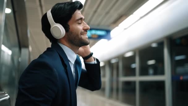 Homem de negócios inteligente ouvindo música por fone de ouvido enquanto espera trem na estação de trem com fundo turvo. Gerente de projeto qualificado desfrutar ouvir relaxar som enquanto segurando o telefone móvel. Exultante. - Filmagem, Vídeo