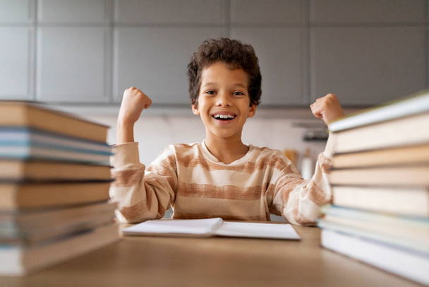 Vidám fekete iskolás fiú, aki otthon ünnepli a sikeres tanulást, boldog fiú, felemelt ököllel és ragyogó mosollyal ül a könyvek mellett, örömöt érez a házi feladat befejezése után - Fotó, kép