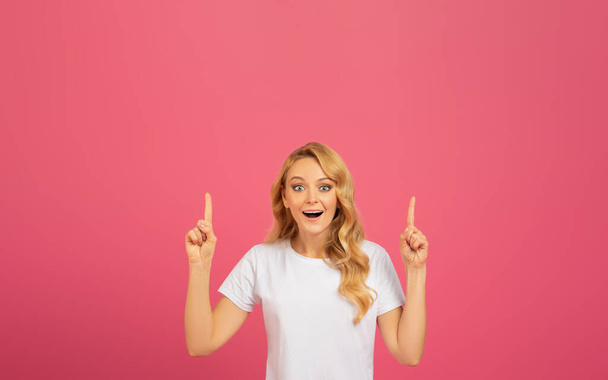 Porträt einer fröhlichen schönen blonden Dame, die mit aufgeregtem Gesichtsausdruck die Finger in die Höhe zeigt, vor rosa Hintergrund im Studio, die mit beiden Händen den freien Raum nach oben zeigt - Foto, Bild