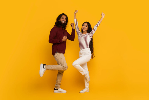 Hombre y mujer enérgicos saltan con brazos elevados y expresiones eufóricas sobre fondo amarillo - Foto, imagen
