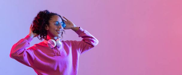 Joyful young woman in pink hoodie, wearing sunglasses and listening to music in white headphones against colorful pink and blue neon background, panorama, free space - Photo, Image
