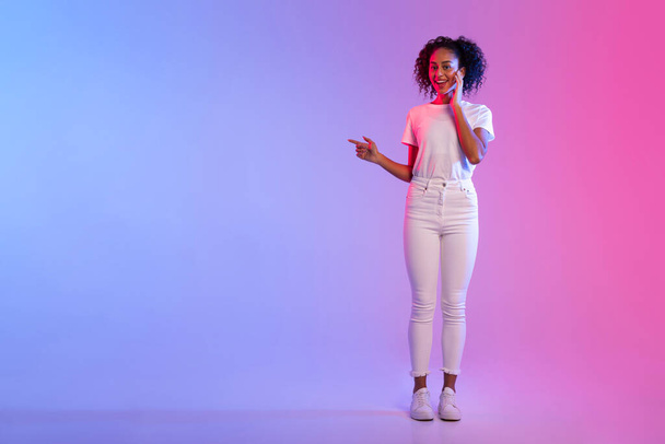 Veselá černá dáma s kudrnatými vlasy ukazuje na stranu ve volném prostoru a usmívá se, představuje reklamu na neonově modré a růžové gradient pozadí, plné délky - Fotografie, Obrázek