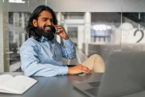 Profissional alegre engajando-se em uma conversa ao telefone enquanto trabalhava com um laptop em sua mesa de escritório - Foto, Imagem