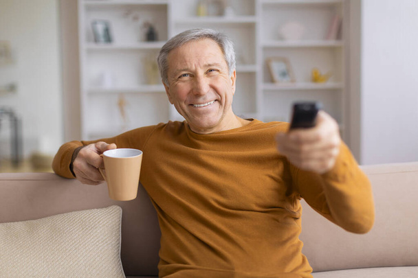 Ένας ηλικιωμένος άνδρας με μια περιστασιακή στολή κρατά μια κούπα καφέ και ένα τηλεκοντρόλ τηλεόρασης, χαμογελώντας καθώς κοιτάζει προς την κάμερα - Φωτογραφία, εικόνα
