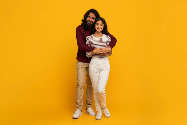Ένα χαμογελαστό ζευγάρι στέκεται κοντά, αγκαλιασμένοι μπροστά σε ένα ζωντανό κίτρινο φόντο, απεικονίζοντας ζεστασιά και στοργή - Φωτογραφία, εικόνα