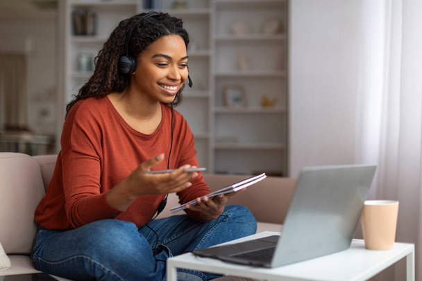 Sorrindo jovem negra vestindo fone de ouvido com microfone se engajando em reunião virtual no laptop em casa, feliz afro-americana tomando notas enquanto trabalhava confortavelmente em sua sala de estar - Foto, Imagem