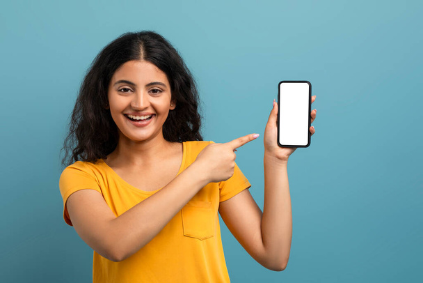 Mujer alegre apuntando a una pantalla de teléfono inteligente en blanco con una gran sonrisa en un fondo verde azulado - Foto, imagen
