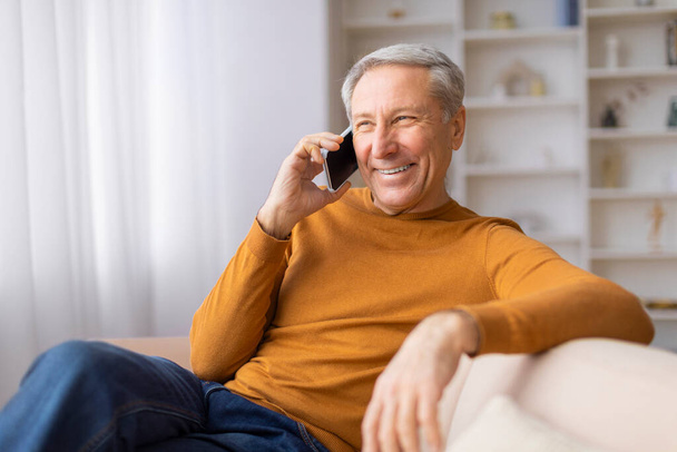 Vidám nyugdíjas ül a kanapén részt vesz egy kellemes beszélgetés az ő smartphone, tükrözve kapcsolat - Fotó, kép