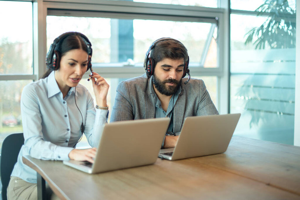 Twee call center agenten praten met klanten tijdens het werken op laptops op kantoor - Foto, afbeelding