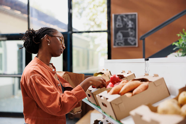 Mujer negra con gafas eligiendo productos cultivados localmente de cajas en estantes en una tienda respetuosa con el medio ambiente. Individuo femenino afroamericano mirando diferentes frutas y verduras frescas. - Foto, imagen