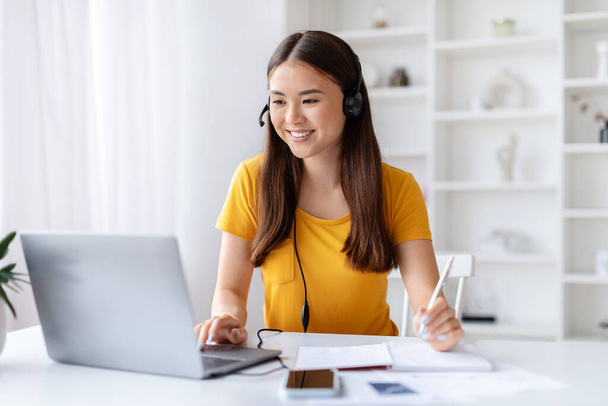 Radosna młoda kobieta nosząca słuchawki wielozadaniowe z piórem i laptopem, robiąca notatki patrząc na ekran komputera, koreańska kobieta pracująca przy biurku w jasnym i zorganizowanym biurze domowym, przestrzeń do kopiowania - Zdjęcie, obraz