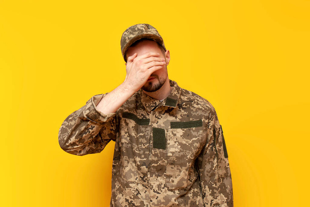 young Ukrainian army soldier in camouflage pixel uniform shows facepalm and failure gesture on yellow isolated background, Ukrainian military cadet fails - Photo, Image